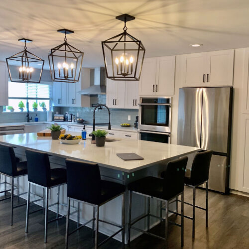 Kitchen featuring large center island surrounded by counter height stools for entertaining guests. White shaker style cabinets with black cabinet and light fixtures.